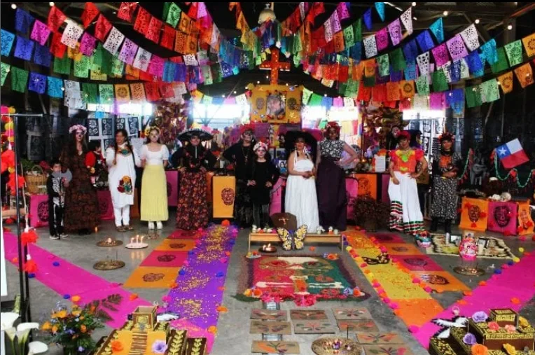 Personas vestidas de catrinas frente al gran altar de muertos en el Galpón Estación, bajo cientos de banderines de papel picado